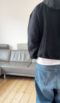 Czarna krótka bluza z kapturem, noszona na białej koszuli, zestawiona z jasnoniebieskimi luźnymi dżinsami z tylnymi kieszeniami, na neutralnym tle.