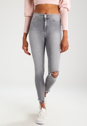 Žena nosící šedé džíny s vysokým pasem, úzkým střihem, roztrženými koleny a třepícími se lemy, doplněné bílými teniskami a krátkým růžovým svetrem.