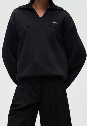 Personne portant un haut noir ample à manches longues avec un petit logo rectangulaire sur la poitrine et un pantalon noir large sur un fond uni.