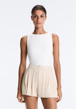 Femme aux cheveux foncés attachés, portant un haut blanc sans manches et un short plissé beige taille haute avec une ceinture assortie.