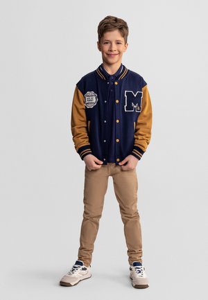 Uśmiechnięty chłopiec stoi, ubrany w granatowo-musztardową kurtkę varsity z naszywkami, beżowe spodnie i białe trampki na jednolitym tle.