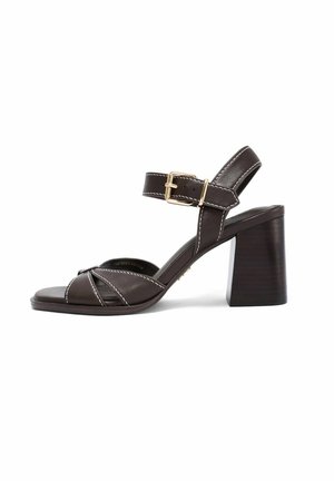 Ciemnobrązowy skórzany sandał na obcasie z paskiem wokół kostki, złotą klamrą, odkrytym noskiem i kwadratowym obcasem na białym tle.