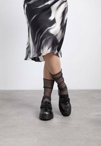 Mocasines negros gruesos con acentos metálicos, combinados con medias negras transparentes y brillantes, llevados con una falda fluidas de patrones en blanco y negro.