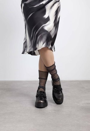 Mocasines negros gruesos con acentos metálicos, combinados con medias negras transparentes y brillantes, llevados con una falda fluidas de patrones en blanco y negro.