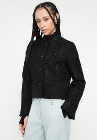 Jachetă neagră scurtă din țesătură texturată, cu design fără guler și închidere cu nasturi, asortată cu pantaloni bleu deschis.