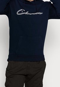 Sudadera azul marino de algodón suave, con un bolsillo frontal tipo canguro y un logo en cursiva blanca en el pecho. Capucha con cordón incluida.