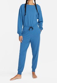Personne portant un sweat-shirt bleu à manches longues et un pantalon de jogging assorti avec des poignets élastiques et un cordon de serrage noir, debout pieds nus.