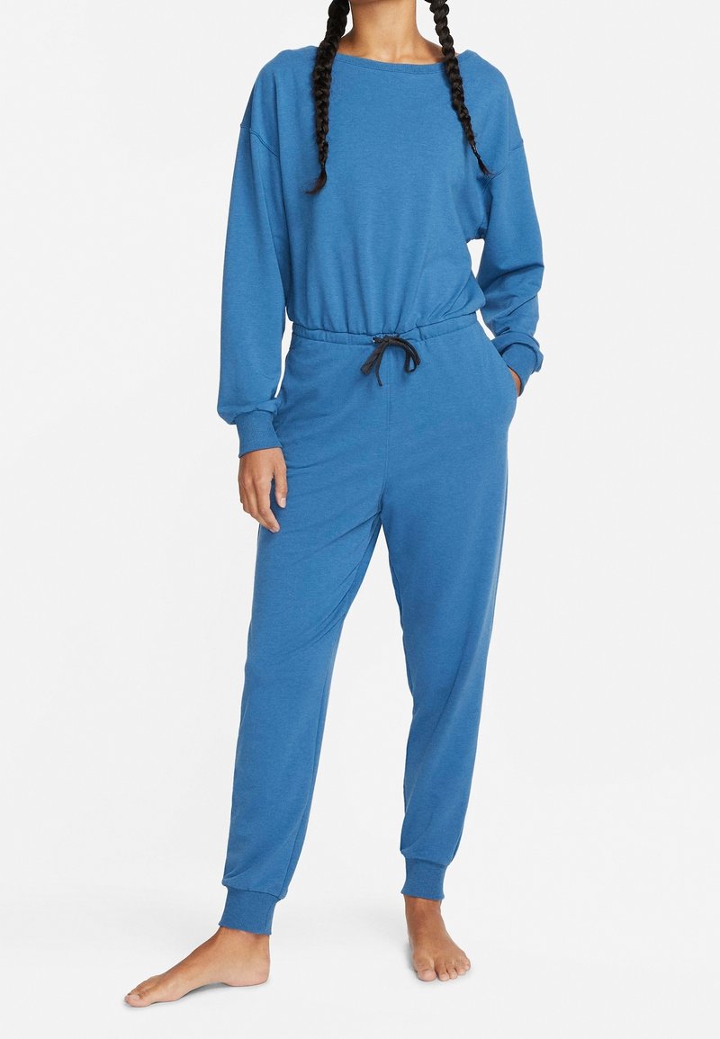 Personne portant un sweat-shirt bleu à manches longues et un pantalon de jogging assorti avec des poignets élastiques et un cordon de serrage noir, debout pieds nus.