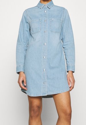 Vaaleansininen farkkupaitamekko, jossa on pitkät hihat, nappikiinnitys edessä, rintataskut ja kaulus, päällä henkilö yksiväristä taustaa vasten.