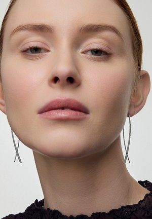 Srebrne kolczyki w kształcie obręczy z krzyżowym wzorem. Gładka skóra modelki kontrastuje z teksturowanym czarnym topem. Neutralne tło.