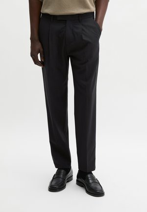 Osoba ubrana w czarne dopasowane spodnie i czarne mokasyny, stojąca z jedną ręką w kieszeni na jednolitym jasnym tle.