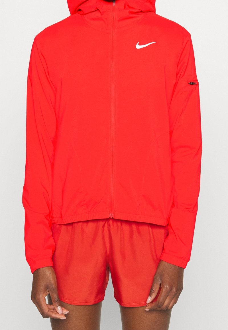 Červená zipová mikina s kapucí, lehký materiál, hladká textura, obsahuje bílé logo Nike na hrudi a zipovou kapsu na rukávu.