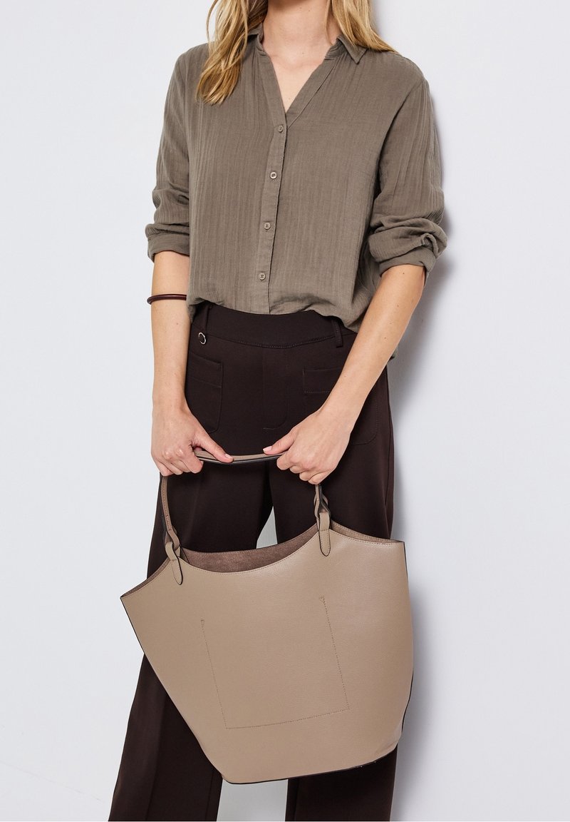 Femme portant une chemise marron à boutons et un pantalon foncé, tenant un grand sac cabas en cuir beige par ses poignées.