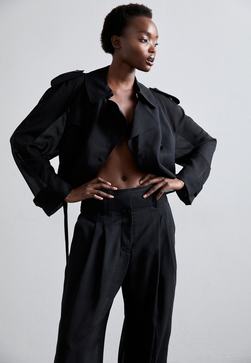 Black trench coat made of sheer fabric with large lapels, paired with high-waisted black trousers. Both garments feature a tailored fit.