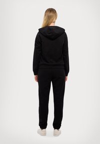 Felpa nera con cappuccio, polsini e vita a coste, abbinata a pantaloni jogger neri coordinati. Il materiale sembra morbido e confortevole.