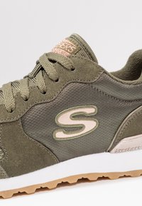 Olijfgroene sneaker met suède en mesh panelen, ronde neus, camouflage textuur en lichtroze logo. Witte tussenzool en bruine rubberen buitenzool.
