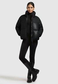 Kvinde iført en sort puffjakke, sorte leggings og sorte knæhøje støvler, stående med hænderne i lommerne foran en ensfarvet baggrund.