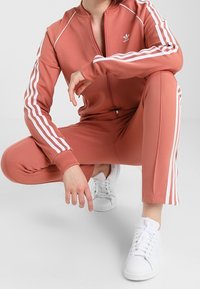 Rose-colored tracksuit made of stretchy fabric, featuring white stripes along the sleeves and pants. Paired with white sneakers.