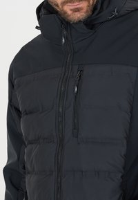 Sort pufferjakke med en glat, mat finish. Har en lynlåslukning foran, justerbar hætte og to lynlåslommer på brystet.
