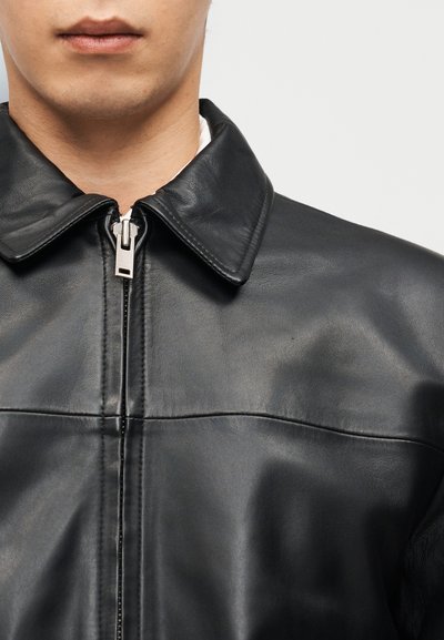 Κοντινό πλάνο στο κάτω μέρος του προσώπου και στον λαιμό ενός ατόμου που φοράει μαύρο δερμάτινο μπουφάν με ασημένιο φερμουάρ και μυτερό γιακά.