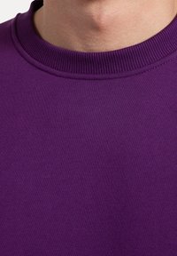 Felpa in cotone viola con scollo a costine. Texture liscia, inquadratura ravvicinata che mette in evidenza il colore uniforme e la consistenza del tessuto.