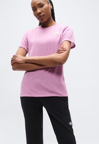 Γυναίκα με σταυρωμένα χέρια, που φοράει ροζ μπλουζάκι North Face και μαύρο παντελόνι North Face, σε απλό λευκό φόντο.