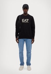 Juodas džemperis su "EA7 Emporio Armani" logotipu, mėlyni džinsai, juoda kepurė ir juoda per petį nešama kuprinė. Sportbačiai yra smėlio spalvos su tekstūruotu akcentu.