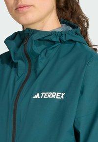 Teal Terrex jacket with a hood, featuring a front zipper and logo in white on the chest. Smooth texture and lightweight fabric.