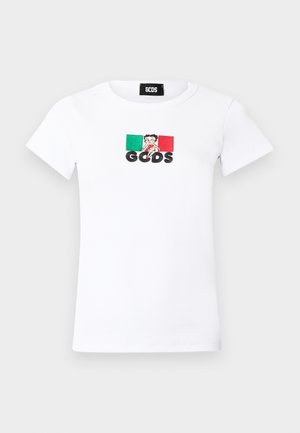 Nő, aki fehér logós pólót, rojtos kék farmer rövidnadrágot, fekete hegyes cipőket visel, és egy fekete kézitáskát tart, egyszerű fehér háttér előtt.