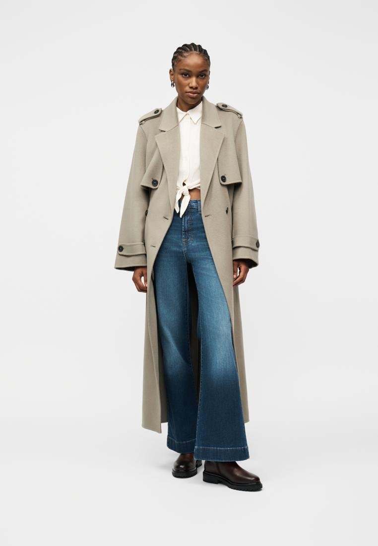 Femme aux cheveux tressés portant un long trench-coat beige, une chemise blanche nouée, un jean bleu à jambes larges et des chaussures noires, debout devant un fond uni.