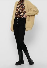 Béžový chlupatý kardigan přes hnědou květinovou halenku, černé džíny s vysokým pasem a pěti knoflíky, a černé kožené kotníkové boty.