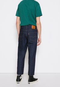 Mörka denimjeans med avslappnad passform, med bakfickor och en brun läderpatch på midjan. Kombinerade med en grön t-shirt och höga sneakers.