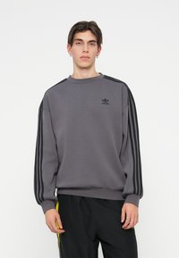 Grå sweatshirt med svart tre-stripes detaljering på ärmarna, ribbad mudd och rund halsringning; har en liten svart logotyp på bröstet.