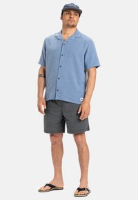 Svetlomodrá košeľa s krátkym rukávom a gombíkmi, šedé šortky a čierne žabky. Košeľa má textúrovanú látku a uvoľnený strih.