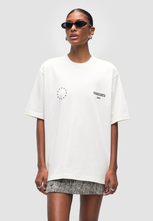 T-shirt bianca in cotone oversize con grafiche circolari e testo sul petto. Il modello indossa occhiali da sole ovali neri e una gonna a fantasia.