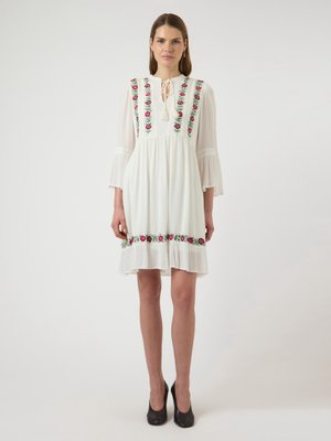 Kobieta stoi, ubrana w białą sukienkę za kolano z różowym kwiatowym haftem na gorsecie i brzegu, z rękawami 3/4 w kształcie dzwonu oraz czarnymi szpilkami.