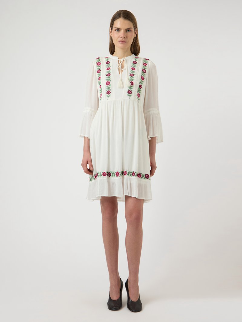 Γυναίκα στέκεται φορώντας ένα λευκό φόρεμα μήκος μέχρι το γόνατο με ροζ κεντήματα με λουλούδια στο μπούστο και το τελείωμα, μανίκια καμπάνα 3/4 και μαύρες γόβες.