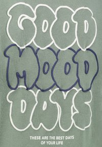 Hellgrüner, strukturierter Stoff mit übergroßer gestickter Schrift: "GUTE HUMOR TAGE" in Weiß und Dunkelblau, mit einer unteren Beschriftung in Weiß.