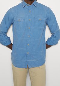 Light blue button-up shirt made of soft fabric, featuring two chest pockets, brown buttons, and rolled-up sleeves.