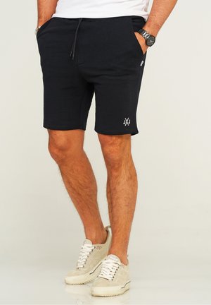 Black cotton shorts with a drawstring waist, side pockets, and a small white logo on the left leg, paired with beige sneakers.