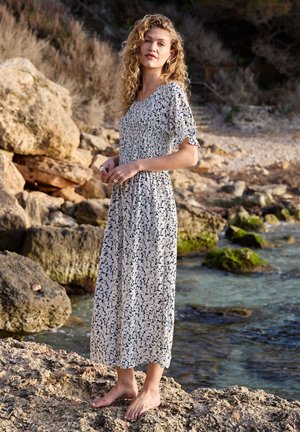 Young woman with curly hair barefoot on rocky shore, wearing a white and black floral midi dress, standing near water and cliffs.