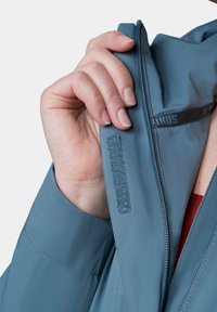 Jasnoniebieska kurtka o gładkiej fakturze, z zamkiem błyskawicznym i paskiem na rzep na wewnętrznej klapie. Ręka trzymająca uchwyt zamka.