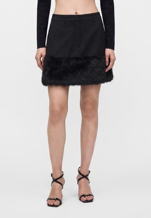 Femme portant une mini-jupe noire avec un ourlet en fausse fourrure et des sandales noires à talons hauts et fines brides sur fond blanc uni.