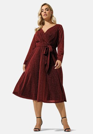 Femme portant une robe portefeuille à manches longues, rouge foncé scintillante, avec une taille nouée et des talons hauts noirs à bride cheville sur un fond uni.