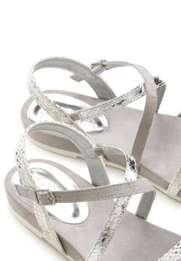 Lichtgrijze sandalen met glanzende zilveren accenten, voorzien van gekruiste bandjes, een zachte textuur op de binnenzool en een lage wiggenhak.