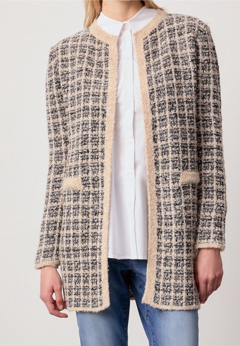 Femme portant un cardigan texturé à carreaux beige et noir par-dessus une chemise blanche boutonnée et un jean bleu, debout devant un fond uni.