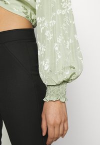 Lys grøn blomstret bluse med puffede ærmer, med en smocked manchet, lavet af et glat stof med en shiny finish. Sorte tætsiddende bukser.