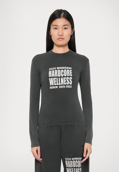 Femeie îmbrăcată cu un tricou și pantaloni de culoare gri închis, cu textul alb „032C WORKSHOP HARDCORE WELLNESS BERLIN 2025-2032”, stând pe un fundal alb.