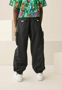 Fekete cargo nadrág elasztikus szegélyekkel, több zsebbel és zöld díszítéssel. Színes grafikai pólóval és fehér sportcipővel viselve.