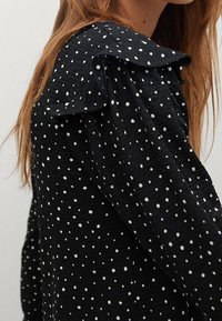 Blouse en tissu noir avec des pois blancs, dotée d'un col volanté et d'une surface texturée. Manches ajustées avec un léger froncé aux épaules.